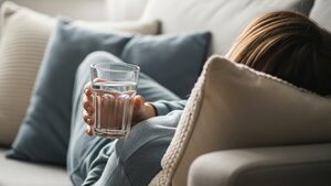 Tired person resting on a couch in soft daylight holding a glass of water.