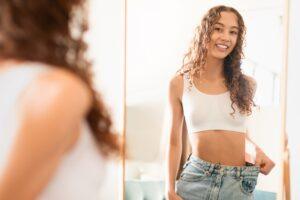 A woman who has lost a significant amount of weight proudly looks in the mirror, holding up her now oversized pants. She wonders how semaglutide works